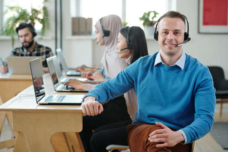Portrait of smiling male call center operator in headset with microphone sitting crossing legs against colleagues working with customer onlineの写真素材