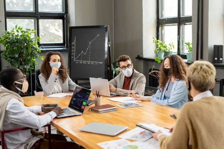Group of colleagues in protective masks discussing working points at meeting in office