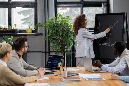 Elegant coach pointing at blackboard while making presentation of financial graph