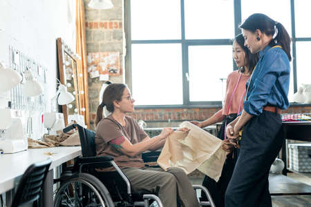 Young fashion designers talking to seamstress in wheelchair in workshopの写真素材