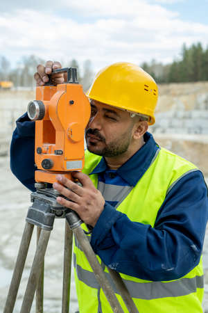 Land surveyor in workwear and hardhat working with geodetic stationの写真素材
