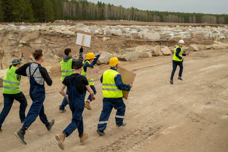 Group of displeased builders with placards running after contractor along construction site