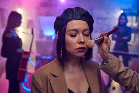 Hand of makeup artist applying powder on face of young woman before recording performance