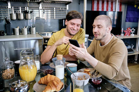 Two guys using smartphone during breakfast by kitchen tableの写真素材