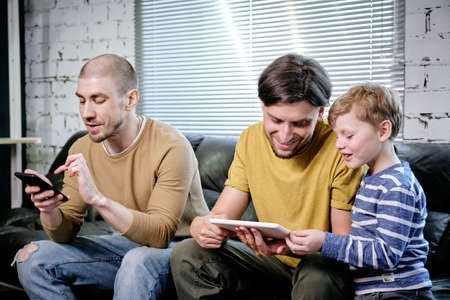 Two young men and little boy using mobile gadgets at leisure