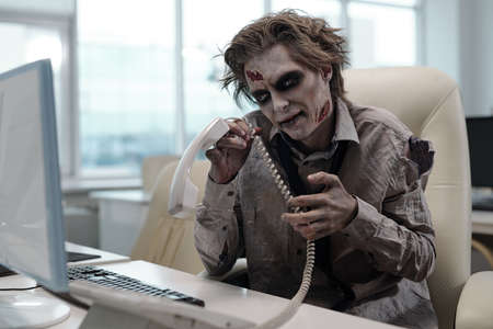 Zombie businessman with phone receiver sitting in front of computerの写真素材
