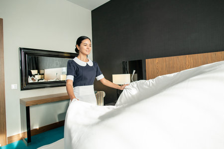 Pretty chambermaid stretching white blanket over bed in hotel room