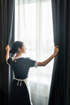 Young chambermaid standing by window in hotel roomの写真素材