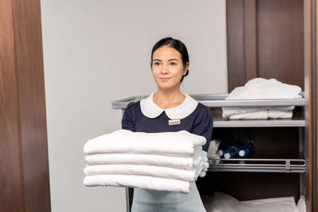Pretty room service staff holding clean sheets while standing by open doorの写真素材