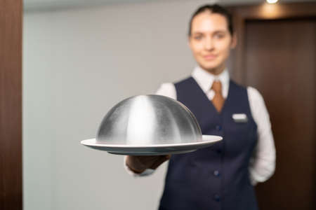 Tray with breakfast held by young female chambermaid in uniform