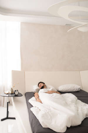 Restful guy sleeping under white sheets in large bedの写真素材