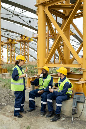 Three intercultural workers in uniform having tea with sandwiches at lunch break