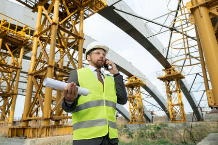 Young engineer in uniform holding smartphone and rolled blueprintの写真素材
