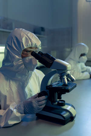 Female microbiologist in coveralls and other protective workwear looking in microscope