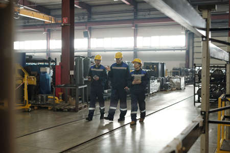 Mature chief engineer in hardhat walking along industrial shop and gesturing hand while giving task...