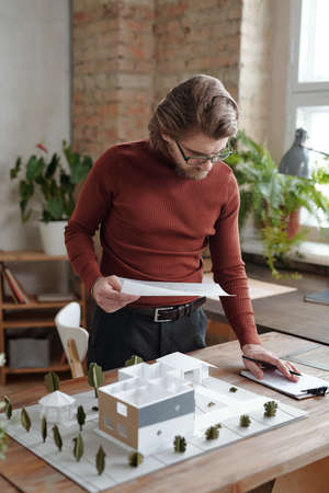 Concentrated young Caucasian building designer in eyeglasses standing at desk and making notes while...