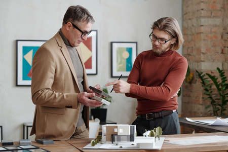Serious mature architect in jacket holding samples and asking question about building design to...
