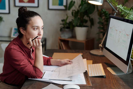 Serious young mixed race woman with hair bun sitting at desk and holding house plan while analyzing...