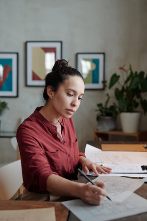 Concentrated young Hispanic woman in burgundy shirt sitting at desk and drawing sketch while...