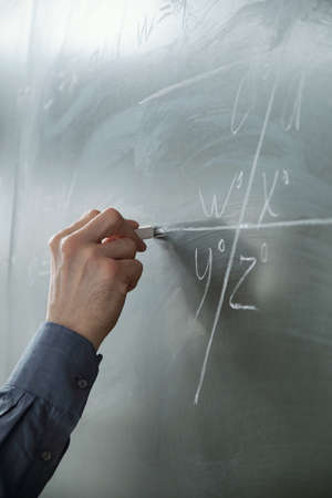 Hand of teacher with chalk by blackboard during explanation at algebra lesson