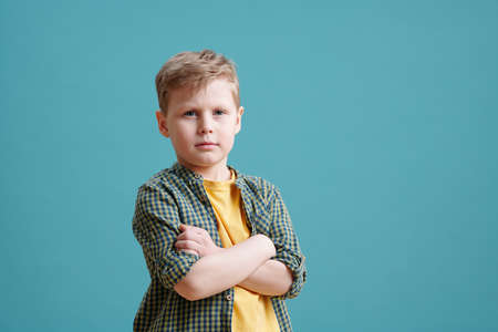 Serious boy keeping his arms crossed by chest while standing on blue backgroundの写真素材