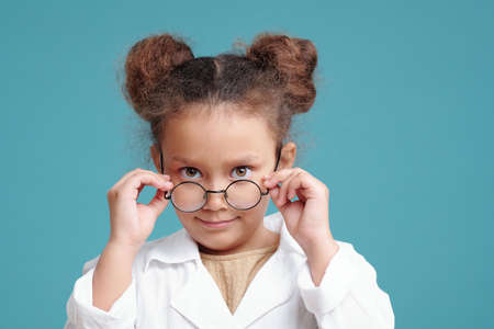 Serious little girl in whitecoat looking at you over eyeglassesの写真素材