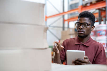 Young African man with digital tablet looking at stack of boxes with goodsの写真素材
