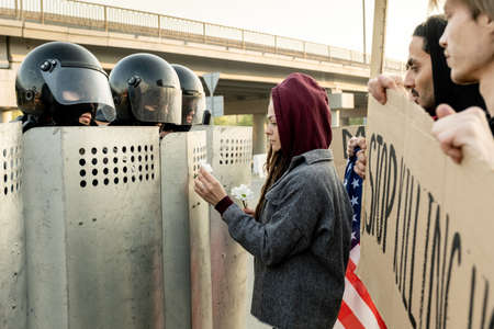 Peace-loving young dreaded woman in jacket giving flowers to police forces moving with riot shields to protesting peopleの写真素材