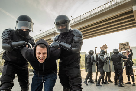 Riot officer in helmets twisting arms of agressive protester in hooded sweater while arresting him...