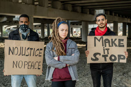 Portrait of serious young interracial people standing with signs under bridge while participating in...