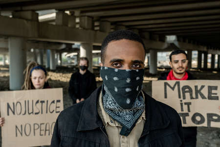 Young Black man in bandana on face standing against protesting people with banners under bridge