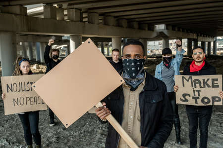 Portrait of serious brutal young Black man with bandana on face standing with banner against team of extremists under bridgeの写真素材