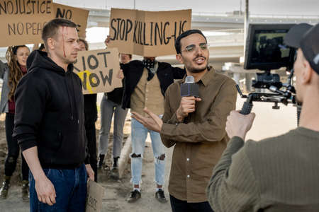 Young mixed race journalist with microphone pointing at protestor with broken head while covering...