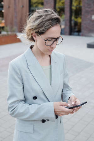 Blond businesswoman in eyeglasses and suit using smartphone outdoors