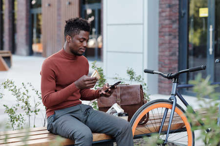 Serious African white collar worker eating sandwich and using smartphone