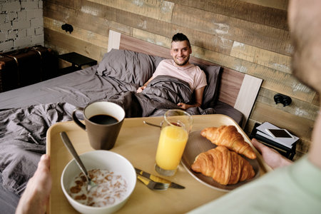 Young man bringing tray with breakfast to his handsome boyfriend in relaxing bedの写真素材