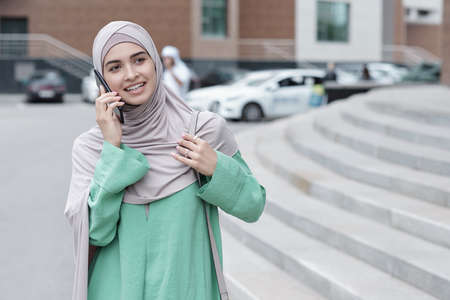 Positive attractive young Muslim woman in hijab standing on city street and chatting on phoneの写真素材