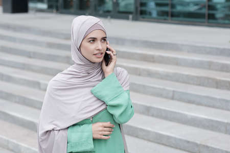 Confident attractive young Islamic woman in green dress standing at office building and talking by phoneの写真素材
