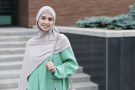 Portrait of positive modern young Islamic businesswoman holding handle of shoulder bag against contemporary office buildingの写真素材
