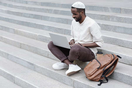 Cheerful handsome young Black man in kufi sitting with laptop between knees and watching webinar outdoorsの写真素材