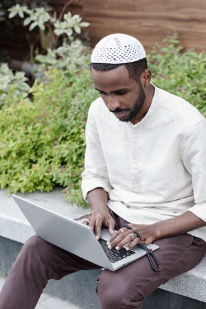 Concentrated busy young Muslim man in kufi cap sitting on curb and working with laptopの写真素材