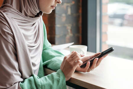 Close-up of calm young woman in hijab sitting at table and answering phone messages In cafeの写真素材