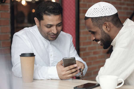Positive young Muslim friends in white shirts sitting in cafe and watching photos on social mediaの写真素材