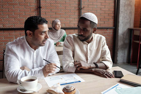 Busy young multi-ethnic businessmen in islamic outfits sitting at table in cafe and discussing chartの写真素材