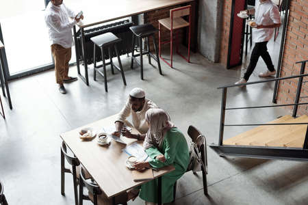 Above view of Islamic people in national clothes sitting at table with papers and working in cafe during lunchの写真素材