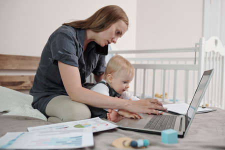 Serious young mother sitting on bed with papers and using laptop while baby touching touchpad of laptopの写真素材