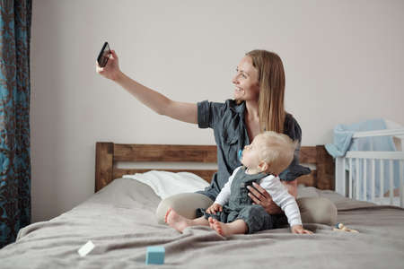 Happy young mother supporting back of son while taking selfie with him on bed