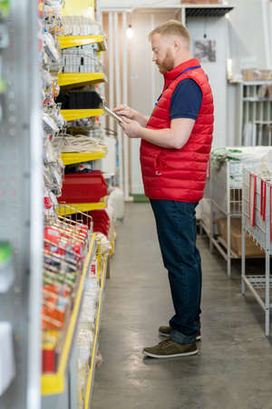 Young man using tablet while making revision of goods on displays