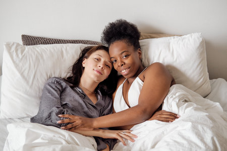 Cute lesbian multiracial couple looking at camera in bedの写真素材