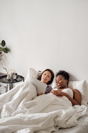 Two young women with tablet relaxing in bed on Sunday morningの写真素材